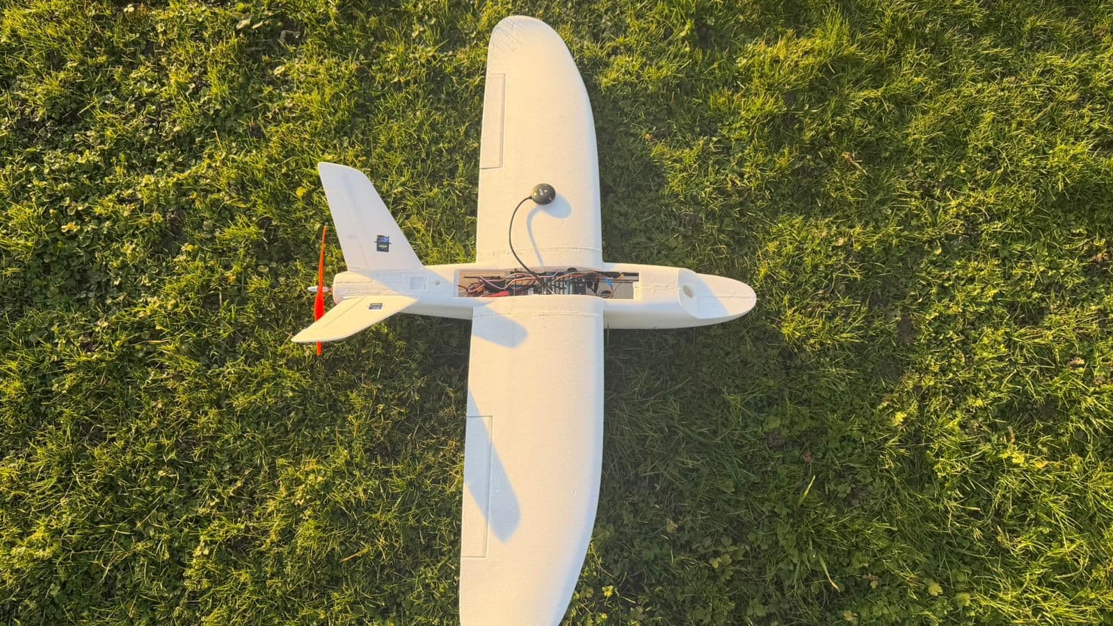 White fixed-wing drone with exposed onboard electronics, lying on grass, seen from above