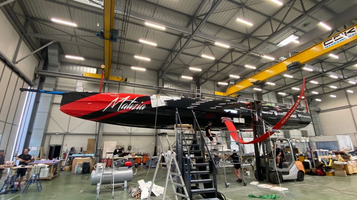 Red and black IMOCA hull in a build hall, surrounded by scaffolding and equipment